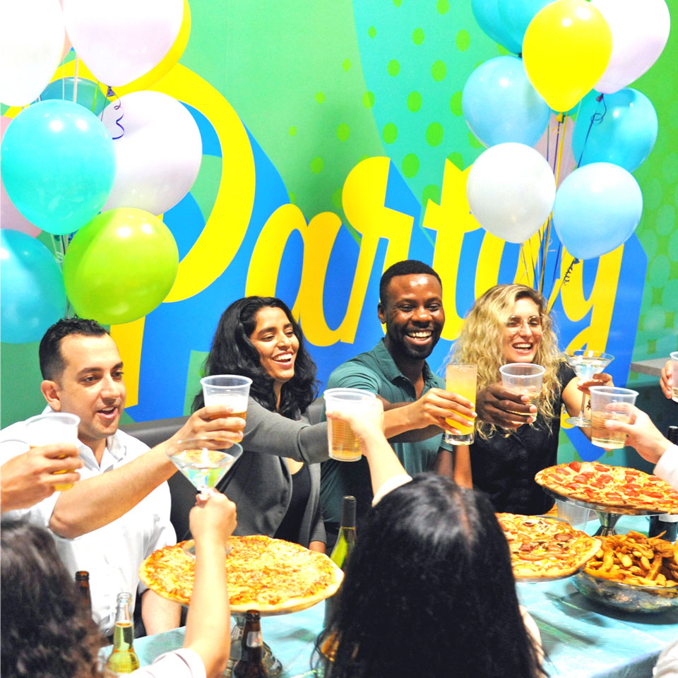 Adults eating pizza and  holding up their drinks at a party with their colleagues.